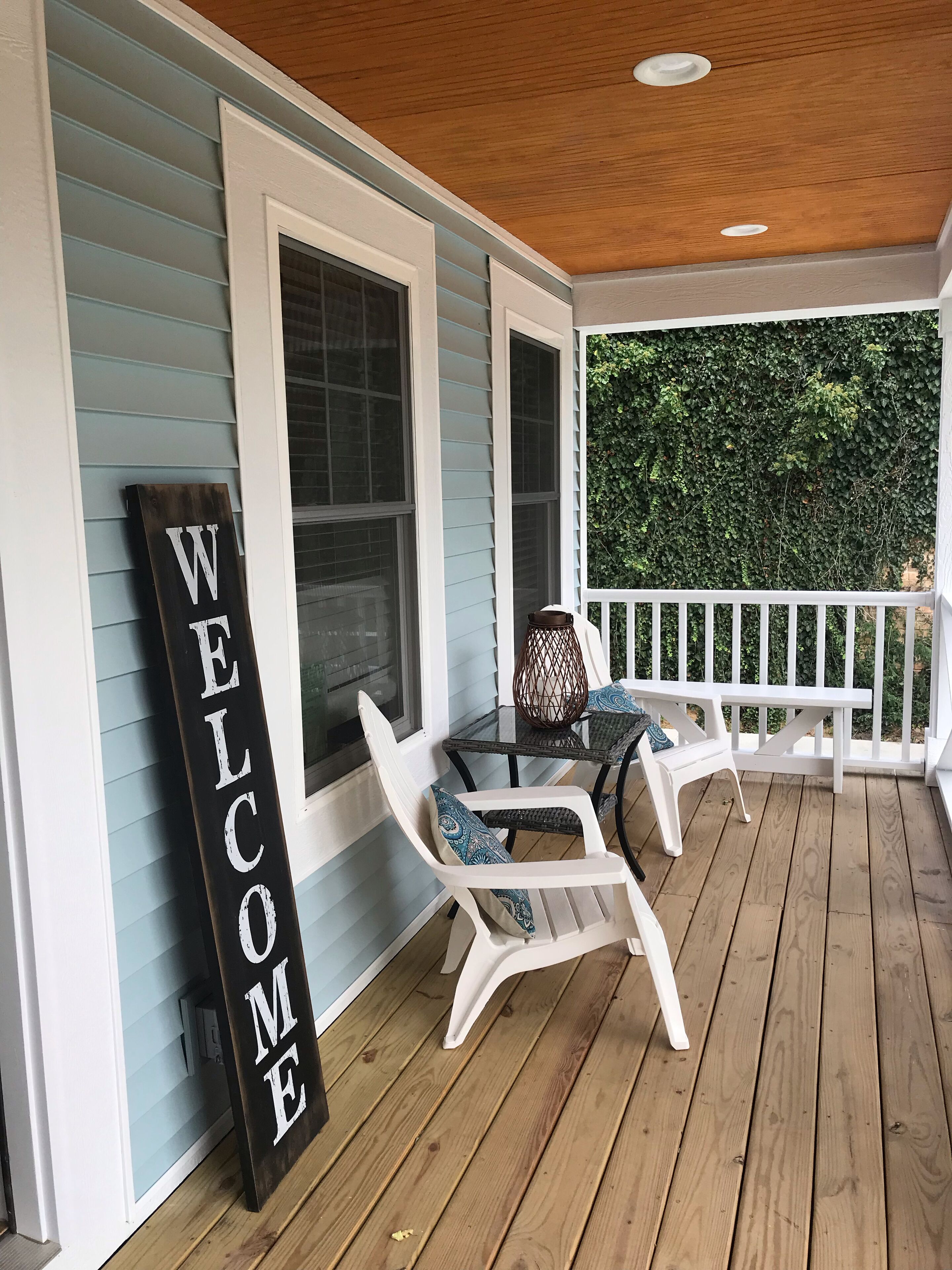 A porch with a 'WELCOME' sign, two white chairs, and a table with a patterned cushion.