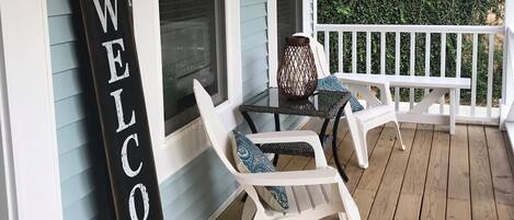 A porch with a 'WELCOME' sign, two white chairs, and a table with a patterned cushion.