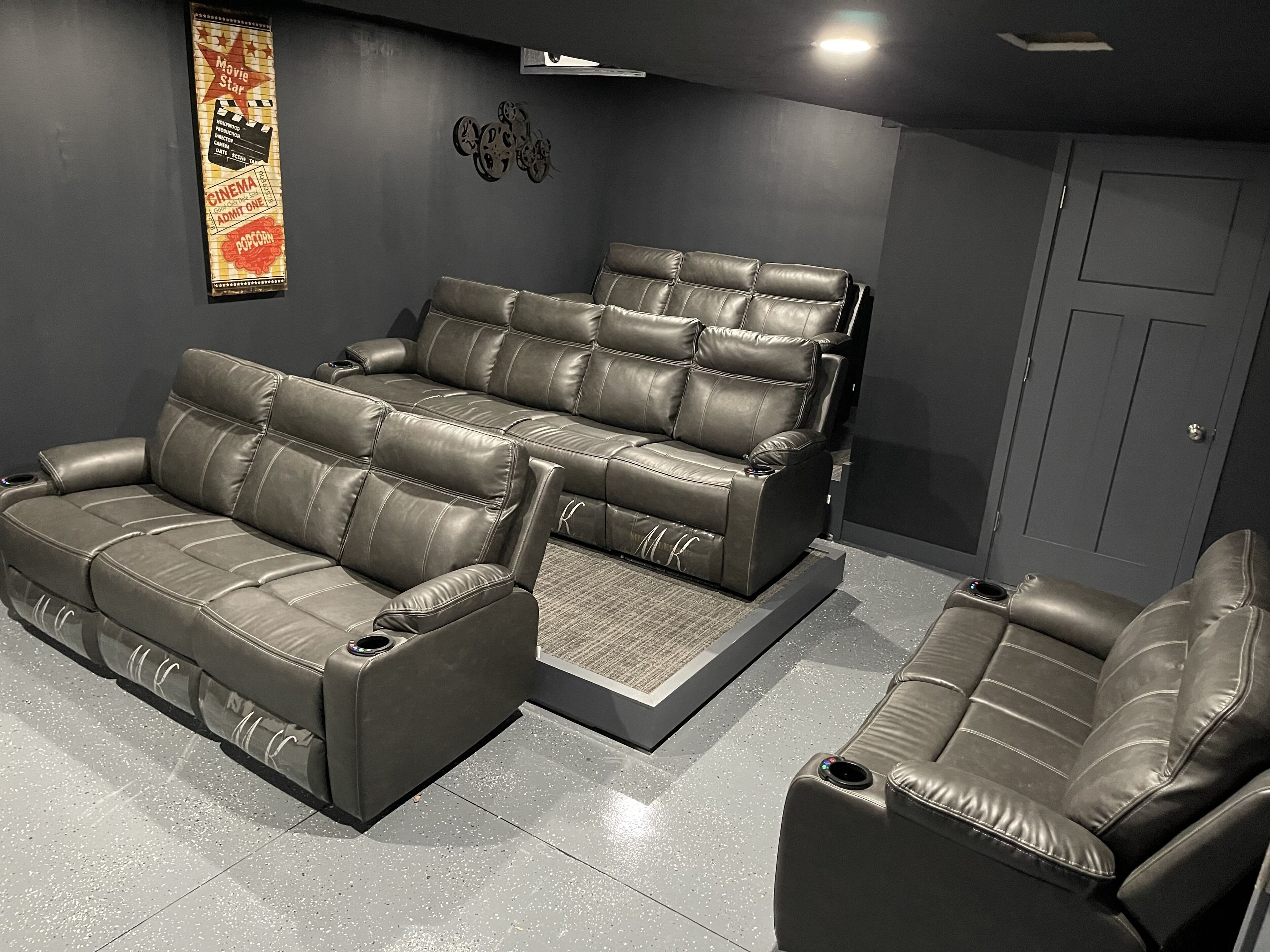 A home theater with a large reclining sofa and a smaller sofa, both in a dark room with a door and a wall-mounted decoration.
