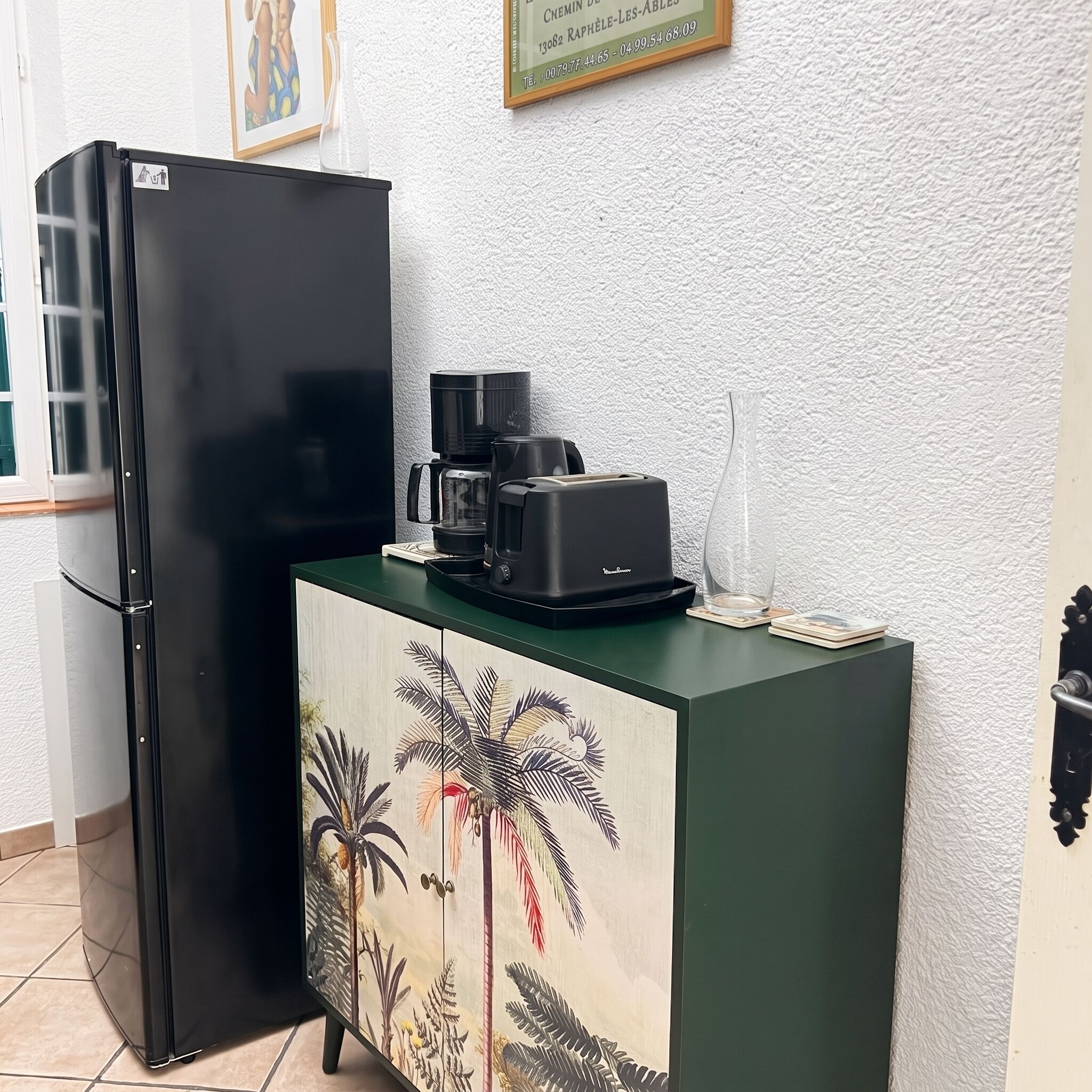 Una cocina con un refrigerador negro, una cafetera y un mueble con diseño de hojas tropicales.
