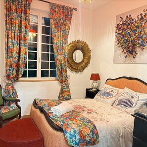 Un dormitorio con una cama, cortinas, un espejo en la pared, una lámpara y un cojín decorativo.
