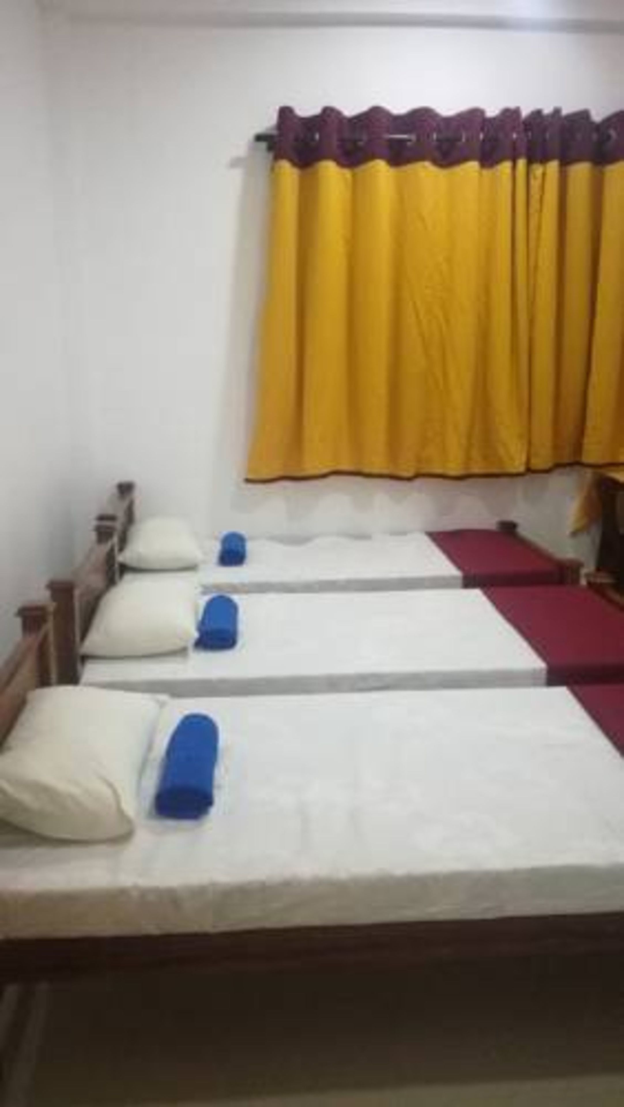 Habitación con tres camas individuales, cada una con una almohada y un objeto azul. Hay una cortina amarilla y un cabecero de madera.
