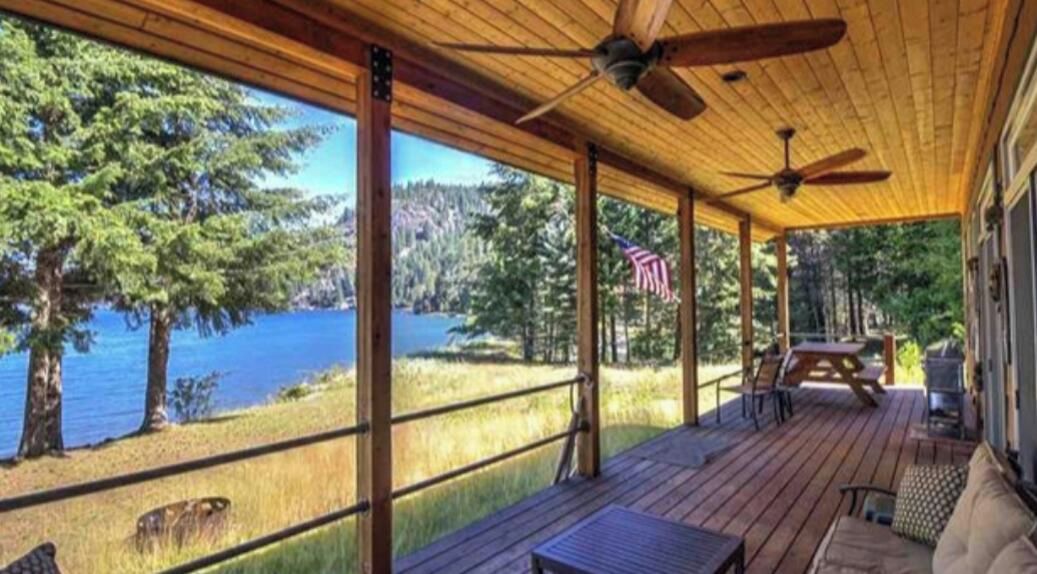 A covered deck with a view of a lake, wooden furniture, and an American flag.