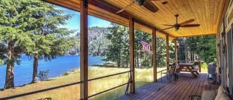 A covered deck with a view of a lake, wooden furniture, and an American flag.