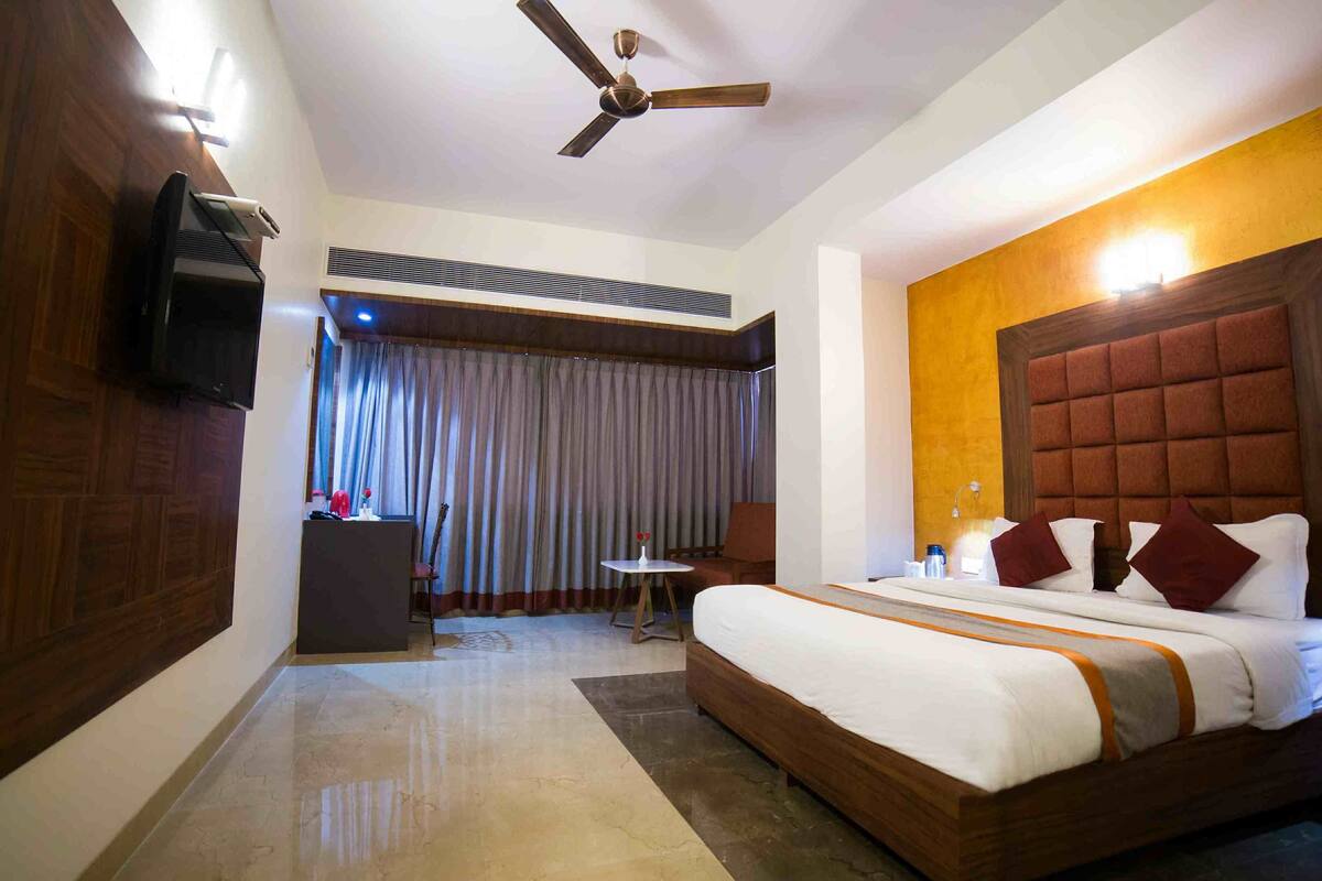 A hotel room with a bed, a desk, a chair, a ceiling fan, and a large window with curtains.