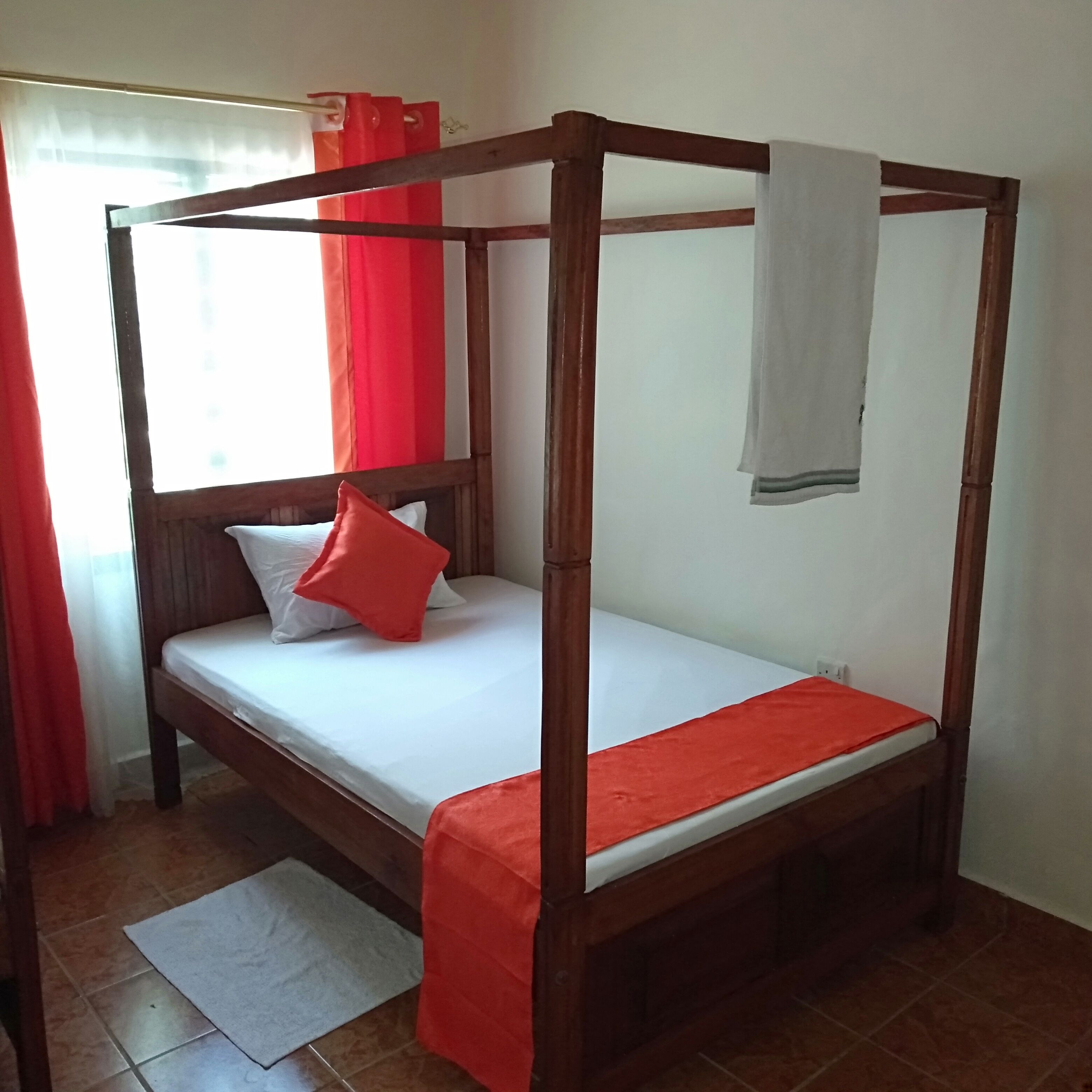 A four-poster bed with a white mattress and red accents, a window with red curtains, and a tiled floor.