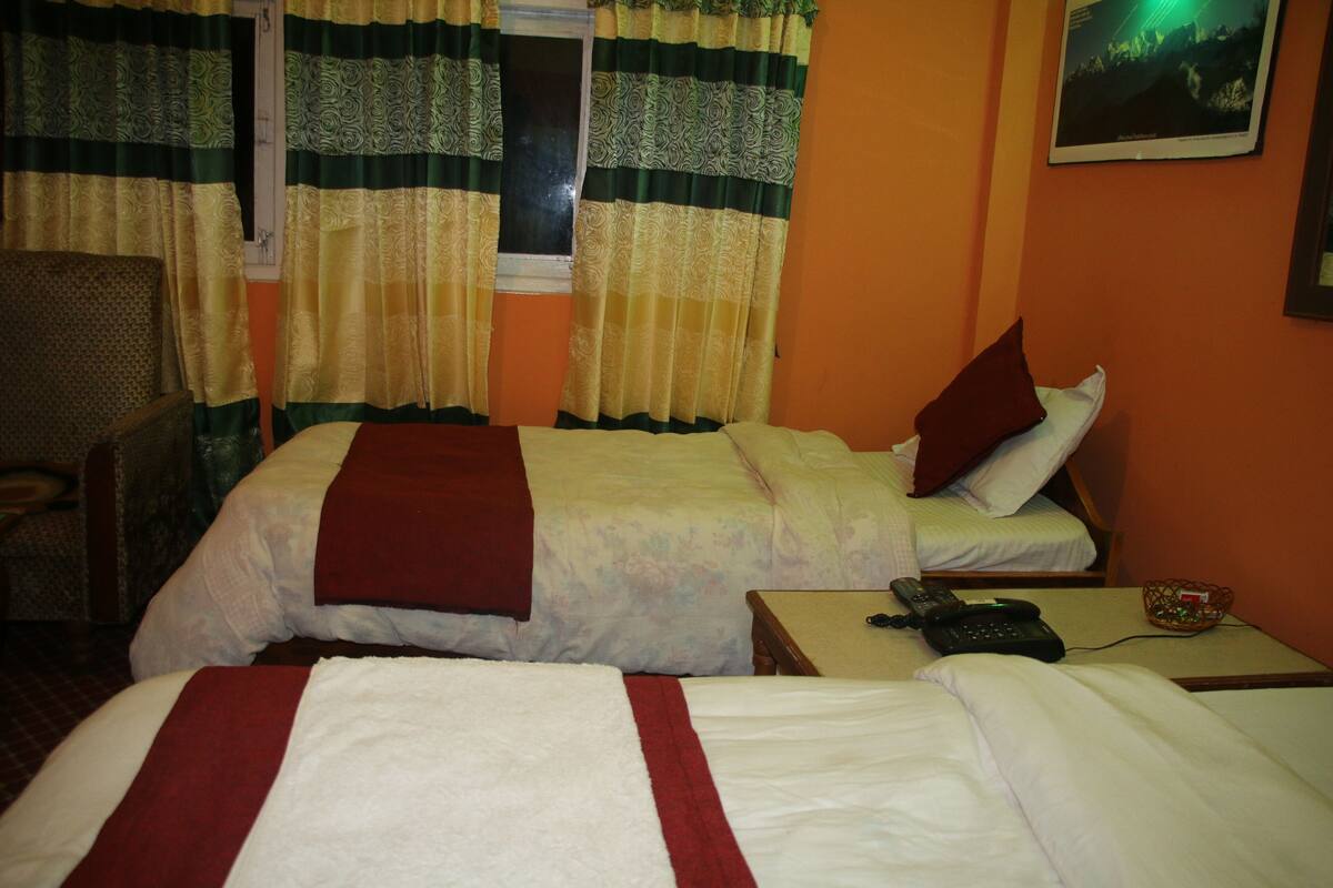 Habitación de hotel con dos camas, una silla, un ventanal con cortinas, un teléfono en la pared y un cuadro enmarcado.