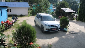 A white SUV parked in a rural setting with a dirt path, surrounded by potted plants and a small blue building. - Taplejung Hotel Pathivara (Taplejung)