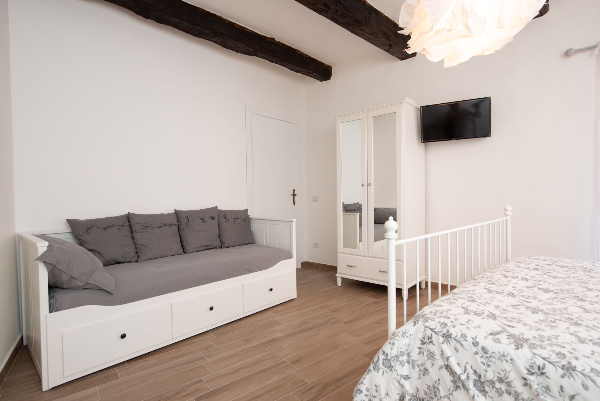 Ein Schlafzimmer mit einem weißen Sofa, einem Bett mit floralem Überwurf, einem Kleiderschrank und einem an der Wand befestigten Fernseher.
