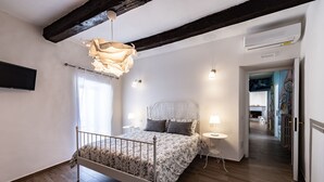 A bedroom with a wooden floor, a bed with a floral bedspread, a hanging light fixture, and a white wall.