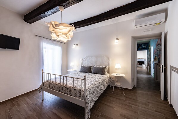Un dormitorio con piso de madera, una cama con colcha floral, una lámpara colgante y una pared blanca.