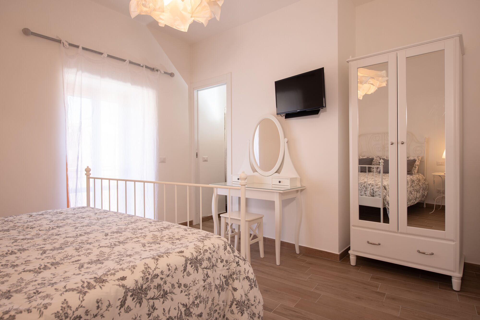 Een slaapkamer met een bed, een kaptafel met spiegel, een kledingkast en een flatscreen televisie aan de muur.