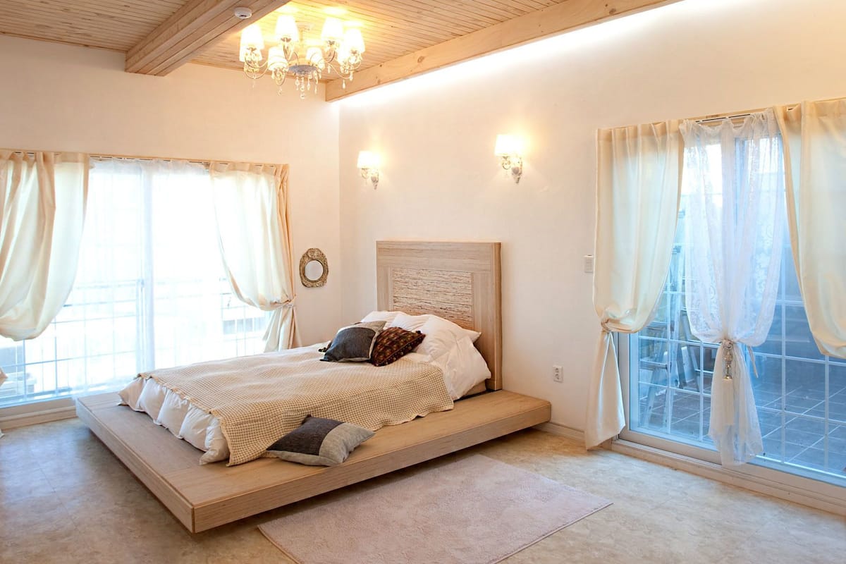 A bedroom with a bed, two windows with curtains, a chandelier, and a small wooden stool with flowers.