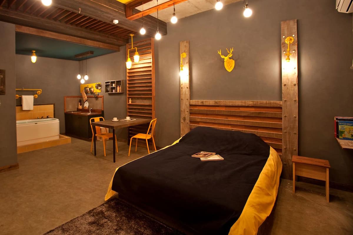 A bedroom with a bed, a desk, a chair, a small table, a bookshelf, a mirror, and a wall-mounted shelf.