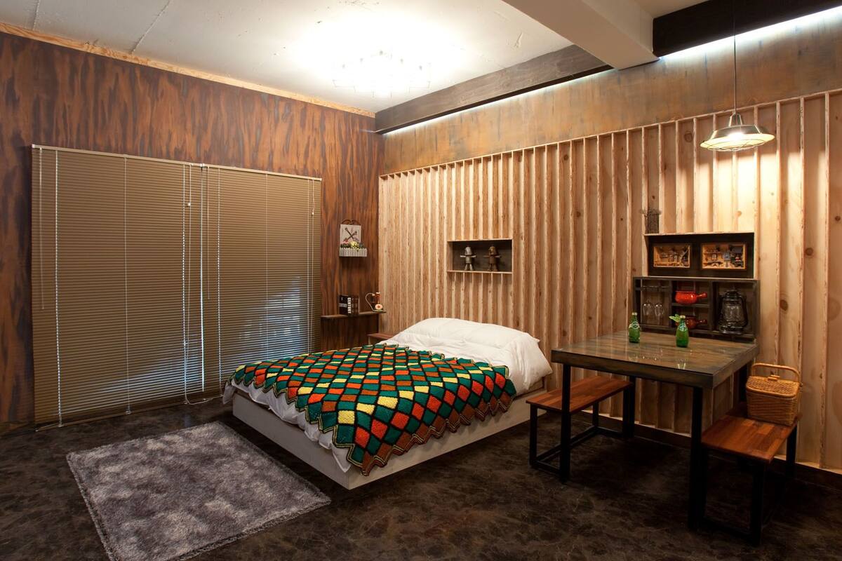 A bedroom with a bed, a small table, a chair, a lamp, and a shelf with decorative items.