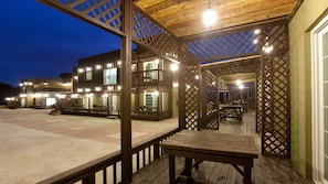 A wooden deck with tables and benches, a covered area with lattice panels, and a building with multiple windows and lights in the background. - Monopoly Pension (Gyeongju)