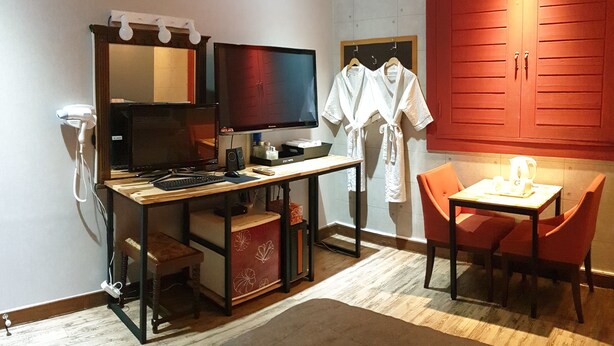 Una camera d'albergo con una scrivania, un computer e una TV. Sono presenti accappatoi appesi al muro e un armadietto rosso.