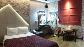 Camera d'albergo con un letto, una toeletta con specchio e vestiti, un tavolo con una ventola e un armadietto rosso.