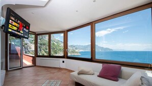A spacious room with a large window offering a sea view, a flat-screen TV mounted on the wall, and a cozy seating area with cushions. - Il Sogno Costa d’Amalfi - Sea View Apartment (Conca dei Marini)