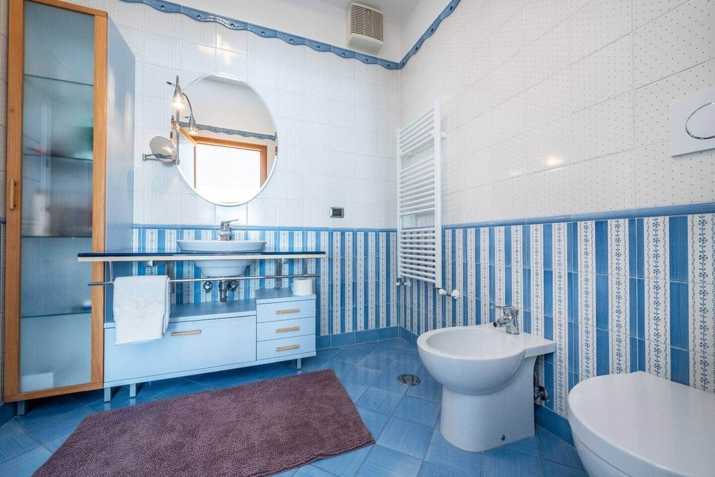 A bathroom with a glass shower enclosure, a sink with a mirror, a bidet, a towel rack, and a patterned tile backsplash.