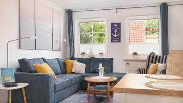 Ein modernes Wohnzimmer mit einem grauen Sofa, einem runden Holztisch und Blick ins Freie durch große Fenster.