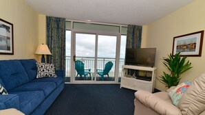 A hotel room with a blue sofa, a beige sofa, a television on a white stand, a plant, and a balcony with chairs. - Bay Watch Resort 419 - Beautiful North Myrtle Beach Oceanfront 1 Bedroom 1 Bath (North Myrtle Beach)