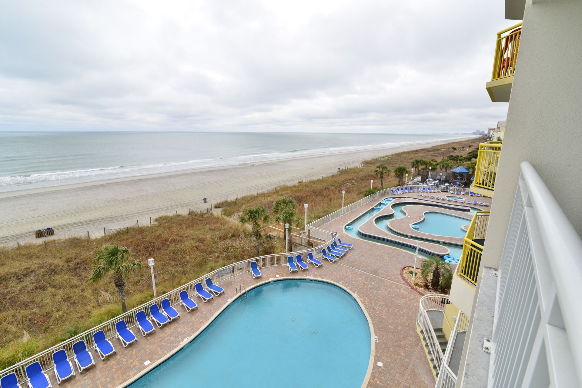 A swimming pool with blue lounge chairs, a beach view, and a building with a yellow railing.