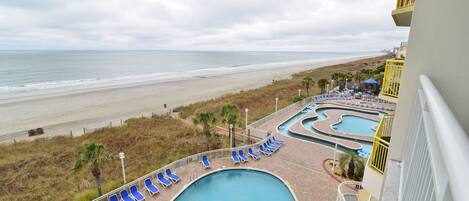 A swimming pool with blue lounge chairs, a beach view, and a building with a yellow railing.