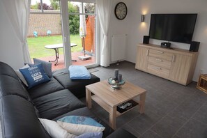 A living room with a black leather sofa, a wooden coffee table, a TV on a wooden cabinet, and a view of the garden through glass doors. - Vacation cottage Nixe, Germany (Wittmund)