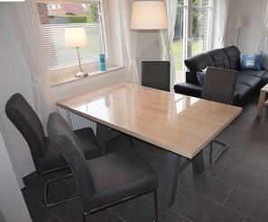 A modern living room with a dining table and chairs, a sofa, and a window with blinds. - Vacation cottage Nixe, Germany (Wittmund)