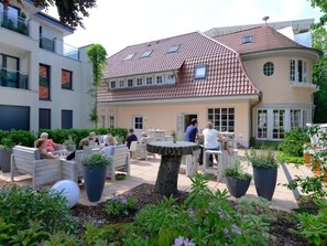 Um pátio com mesas ao ar livre, uma árvore central e um edifício com telhado de azulejos.