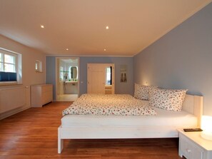 Quarto com cama, mesa de cabeceira e apoio para objetos pessoais. Há uma janela no lado esquerdo e uma porta que dá para outro cômodo.