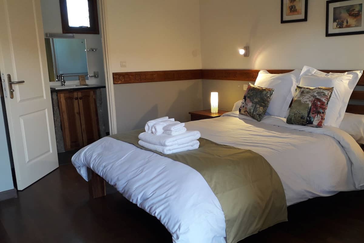 A hotel room with a large bed, a bedside table with a lamp, a nightstand with a lamp, and a bathroom visible through an open door.
