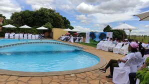 A poolside event with attendees seated under umbrellas, a pool, and a mural in the background.