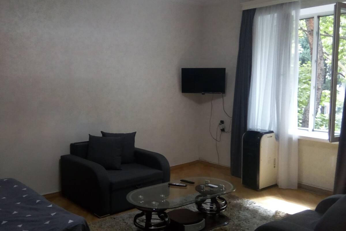 A small, sparsely furnished room with a sofa, a glass coffee table, and a television mounted on the wall.