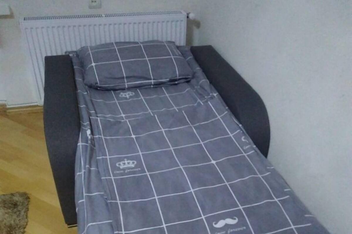 A small, single-bed room with a grey checkered bedspread, a white radiator, and a dark-colored bench or sofa against the wall.