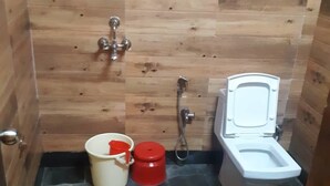 A small, compact bathroom with a wooden wall, a white toilet, a shower, and two buckets. - Banaras Hotel LLP (Kanpur)