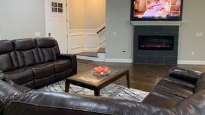 Een woonkamer met een leren bank, een glazen salontafel met fruit, een open haard en een tv die erboven is bevestigd.