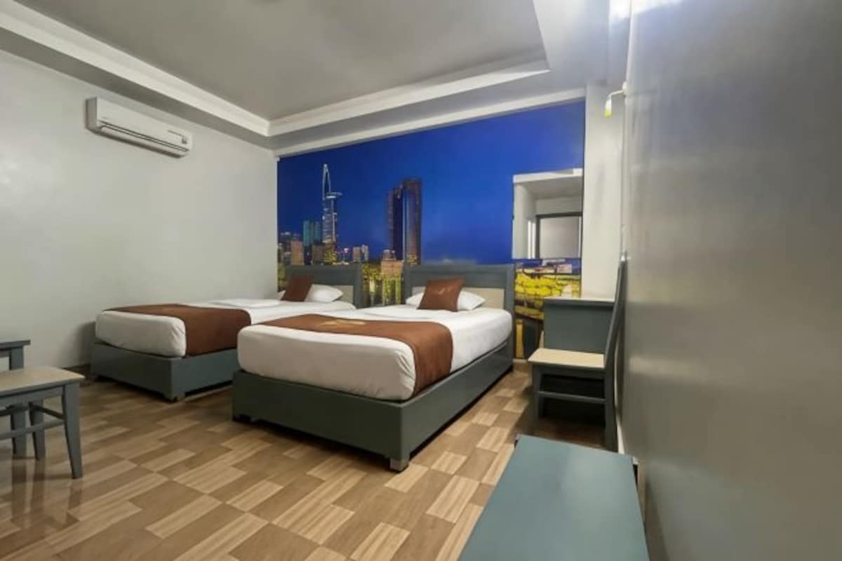 Quarto de hotel com duas camas, uma escrivaninha, uma cadeira, um refrigerador e um mural com vista para a cidade.