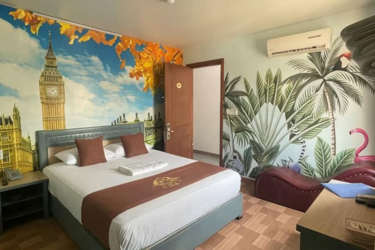 Quarto de hotel com uma cama grande, mesa de cabeceira, escrivaninha, cadeira, um mural na parede e uma porta.