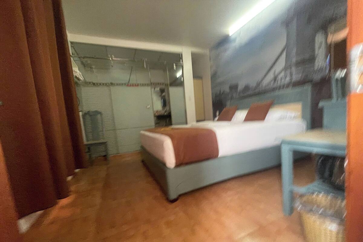 A hotel room with a bed, a small table, and a chair. There is a wardrobe and a mirror in the background.