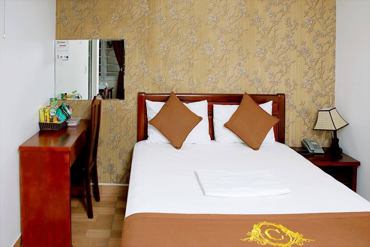 A hotel room with a bed, a desk, a chair, a nightstand with a lamp, and a mirror.