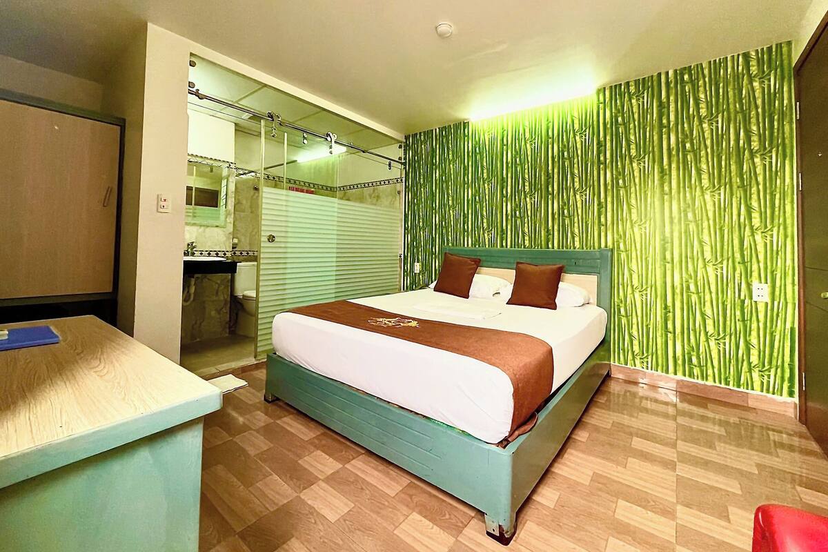 A hotel room with a bed, a desk, a mirror, and a green bamboo-patterned wall.
