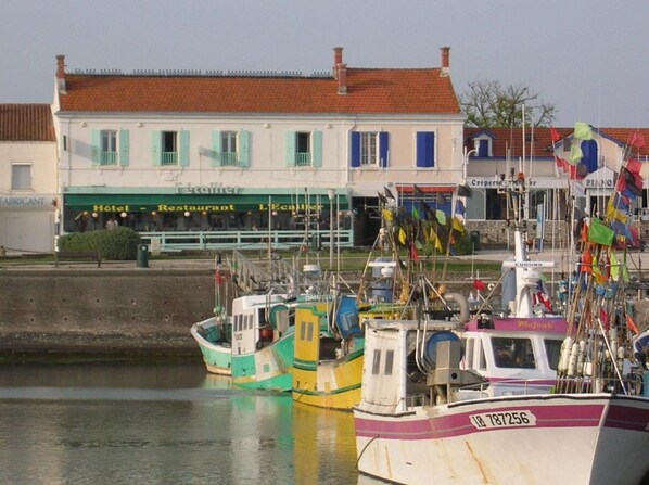 A harbor with several boats docked, a building with a sign reading "Hotel - Restaurant - L'Ecaille", and a red-roofed building in the background. - HĂ´tel Restaurant L'Ecailler (Saint-Pierre-d'Oleron)