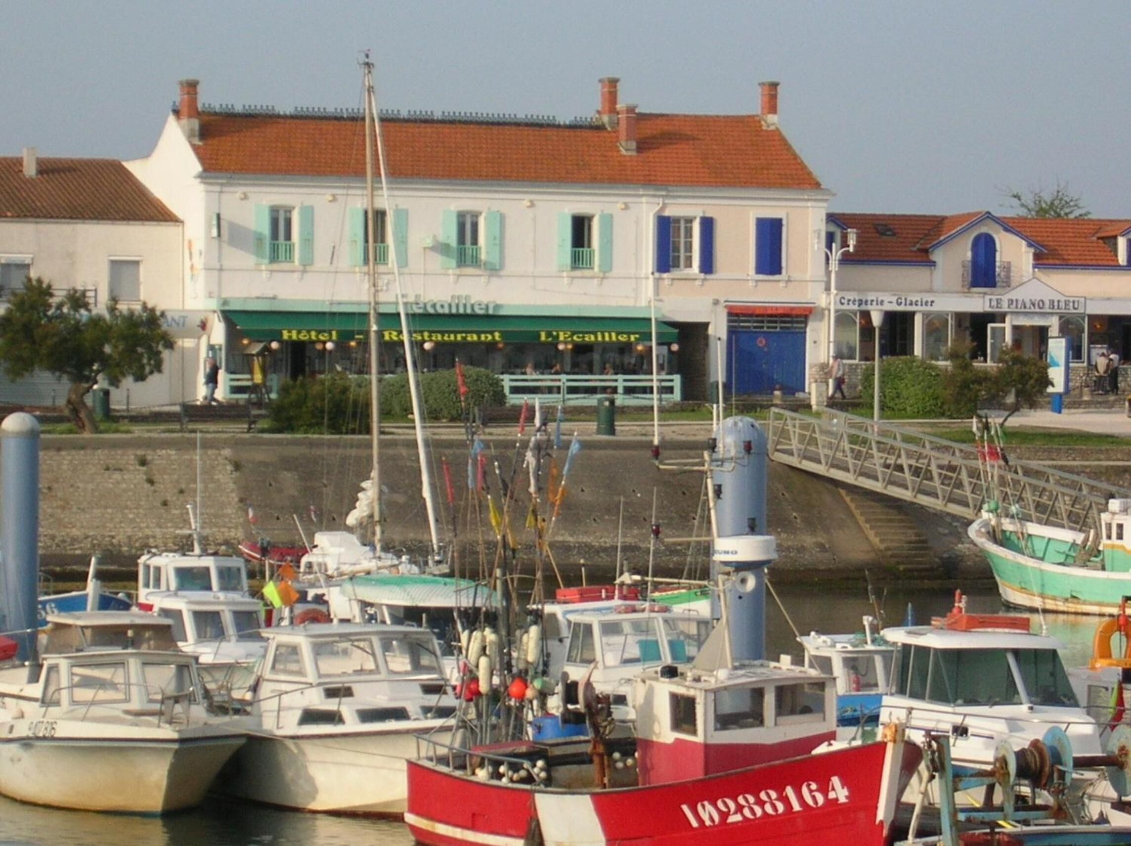 Un port où plusieurs bateaux sont amarrés, un bâtiment sur lequel on peut lire l’inscription « Hôtel-restaurant L’Écaille », et un pont menant à un autre bâtiment sur lequel on peut lire l’inscription « Le Piano Bleu ».