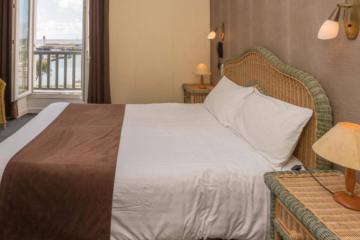 A hotel room with a bed, a chair, a nightstand, a lamp, and a view of the sea through a window.