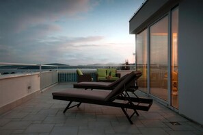 Une terrasse sur le toit, dotée de chaises longues et d’une table, offrant une vue sur une étendue d’eau et sur des collines lointaines.