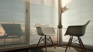A modern room with a glass table, two white chairs, a white vase with flowers, and a view through large windows with blinds. - Sunset Penthouse Apartment with Hot Tub (Zadar)