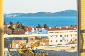 Un balcon avec une table en osier sur laquelle sont disposés des fruits et des boissons, offrant une vue sur un paysage urbain côtier avec ses bâtiments et la mer.