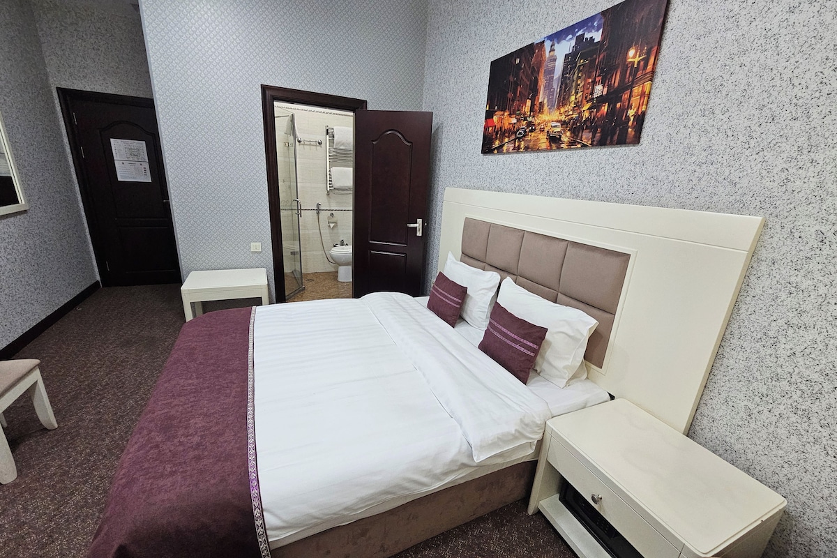 A hotel room with a bed, a nightstand, a small table, a bathroom visible through an open door, and a cityscape painting on the wall.